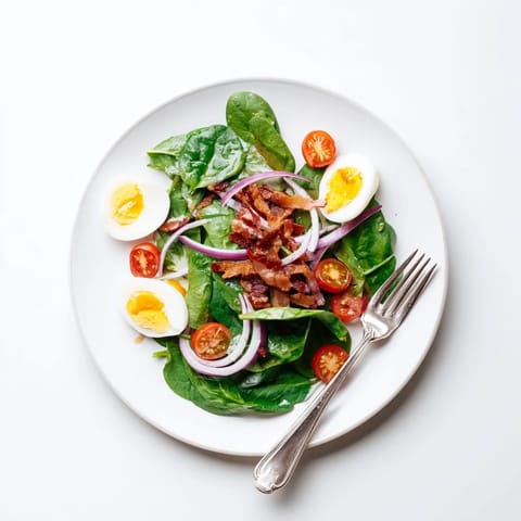 A close-up of a generous Spinach Salad with Bacon, showcasing golden crispy bacon pieces, creamy egg slices, and vibrant red onion on fresh greens.